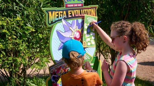 Two children interact with a Pokémon Trading Card Game Mega Evolution display outdoors, writing on a worksheet with trees in the background.